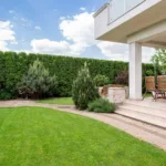 Un angolo sereno di un giardino moderno con pavimentazione in pietra, sedie in legno e una ricca vegetazione.